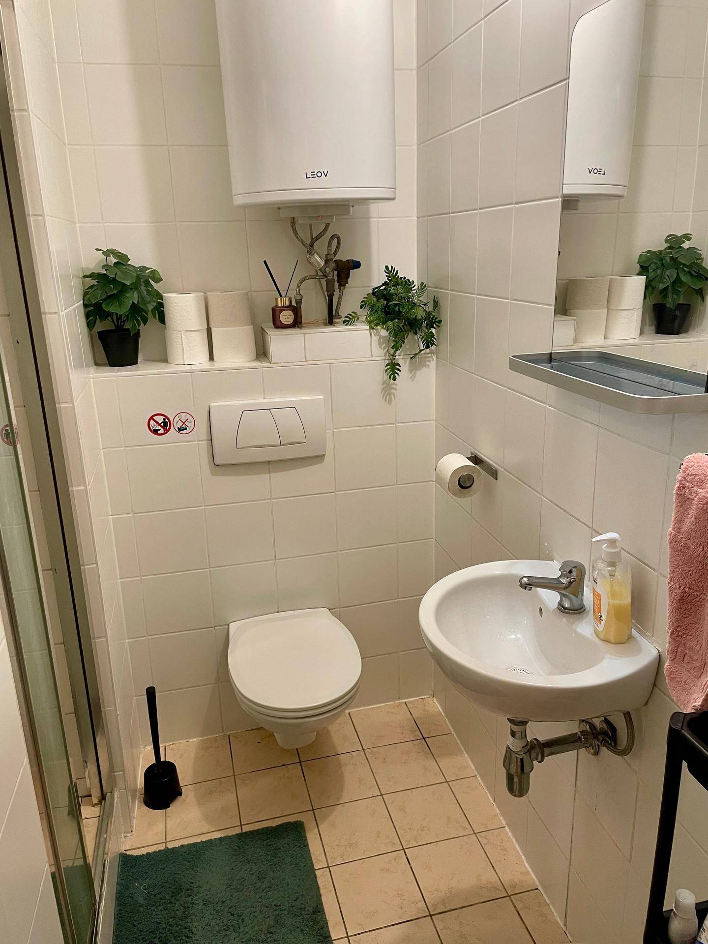 Kleines Badezimmer mit weißen Fliesen, Toilette, Waschbecken und Duschbereich.