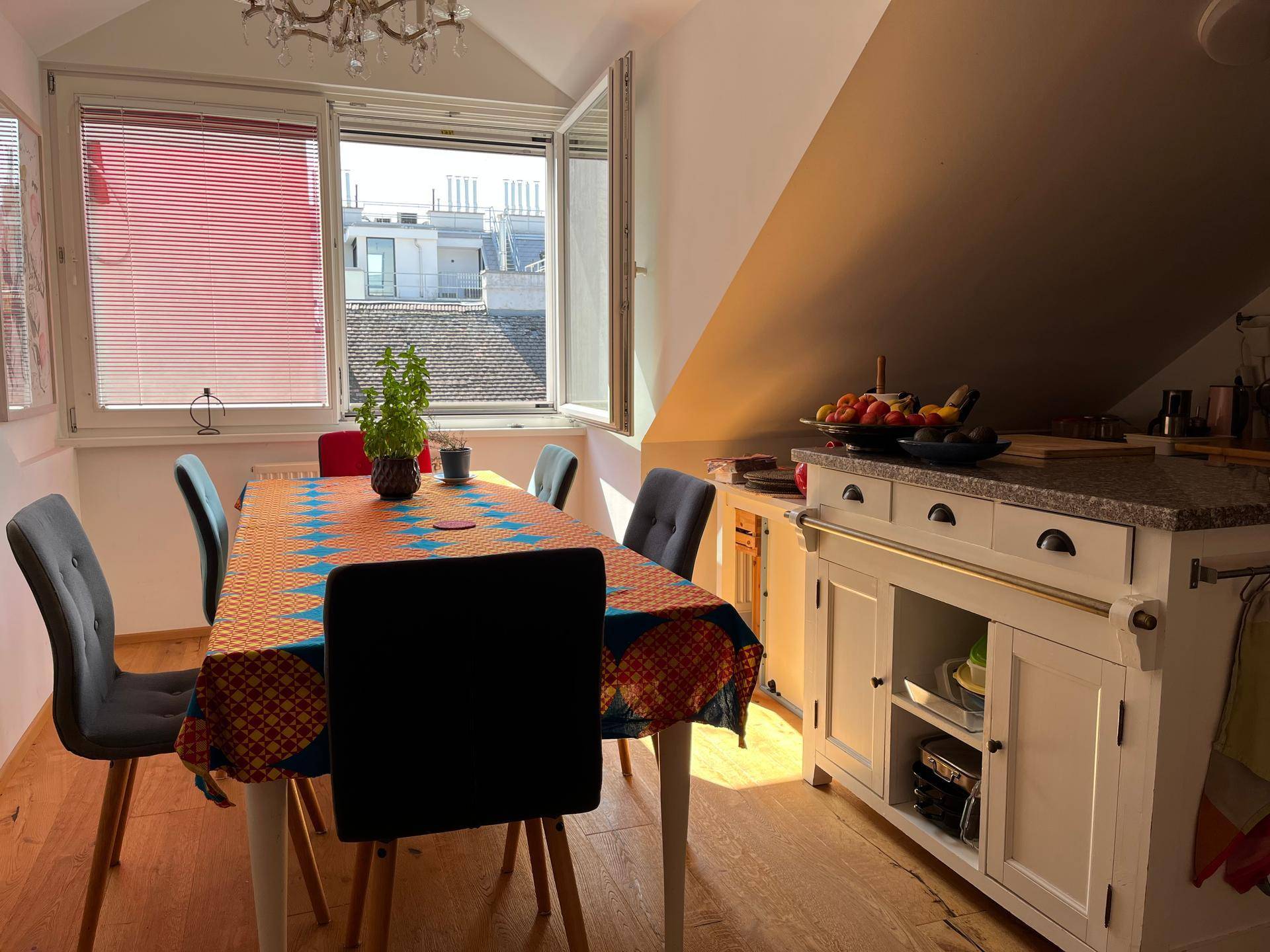 Heller Essbereich mit großem Tisch, farbenfroher Tischdecke und Fenster mit Blick auf die Dächer.