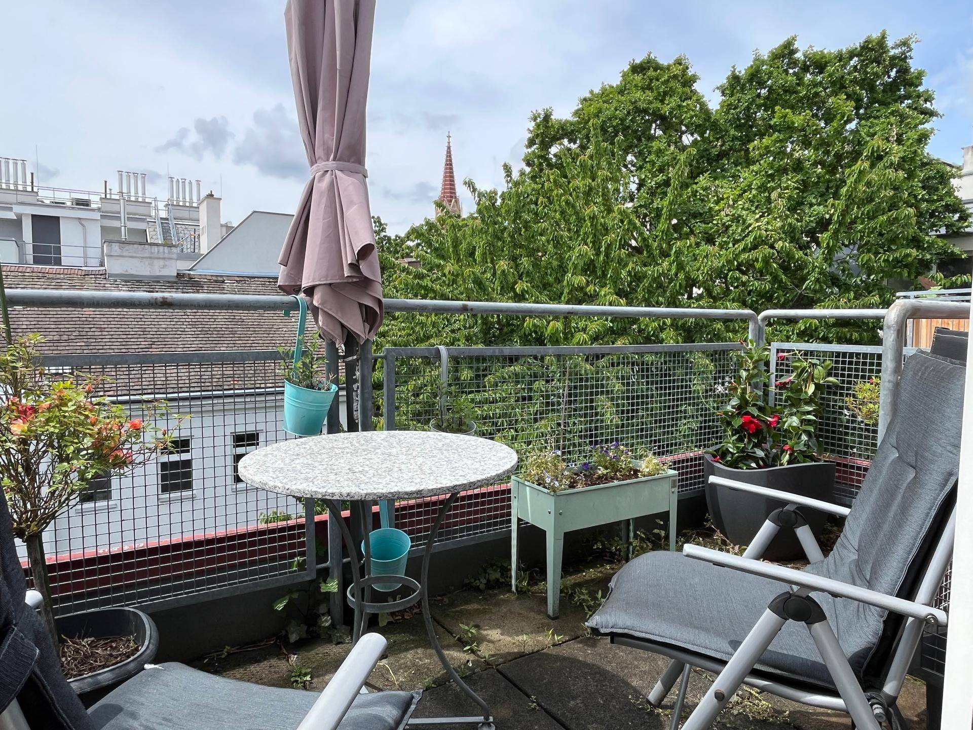 Balkon mit Sitzgelegenheiten, Pflanzen und Blick über die Dächer und grüne Bäume der Stadt.