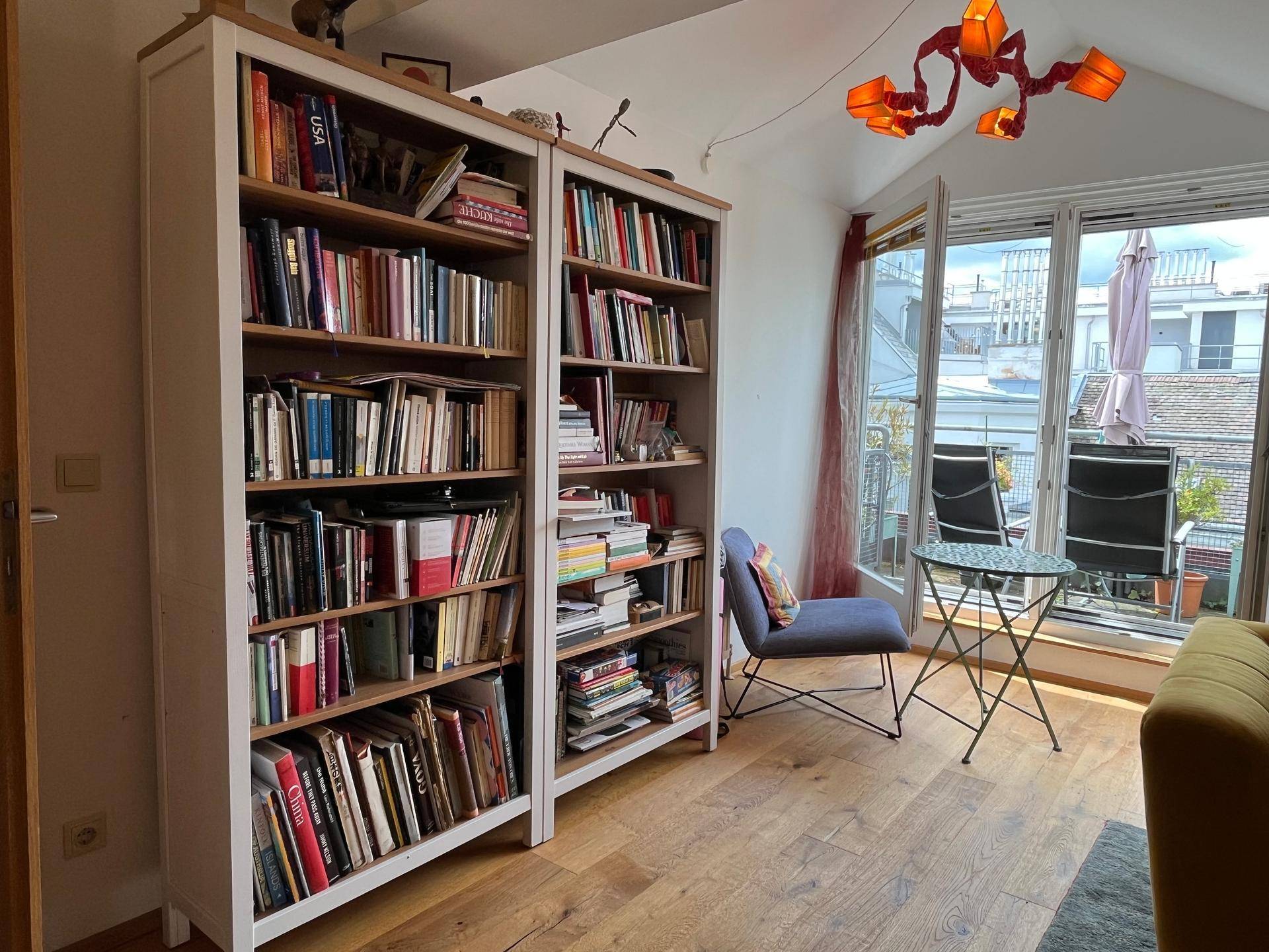 Wohnzimmer mit Bücherregalen, kleinem Sitzbereich und Zugang zum Balkon durch große Fenster.
