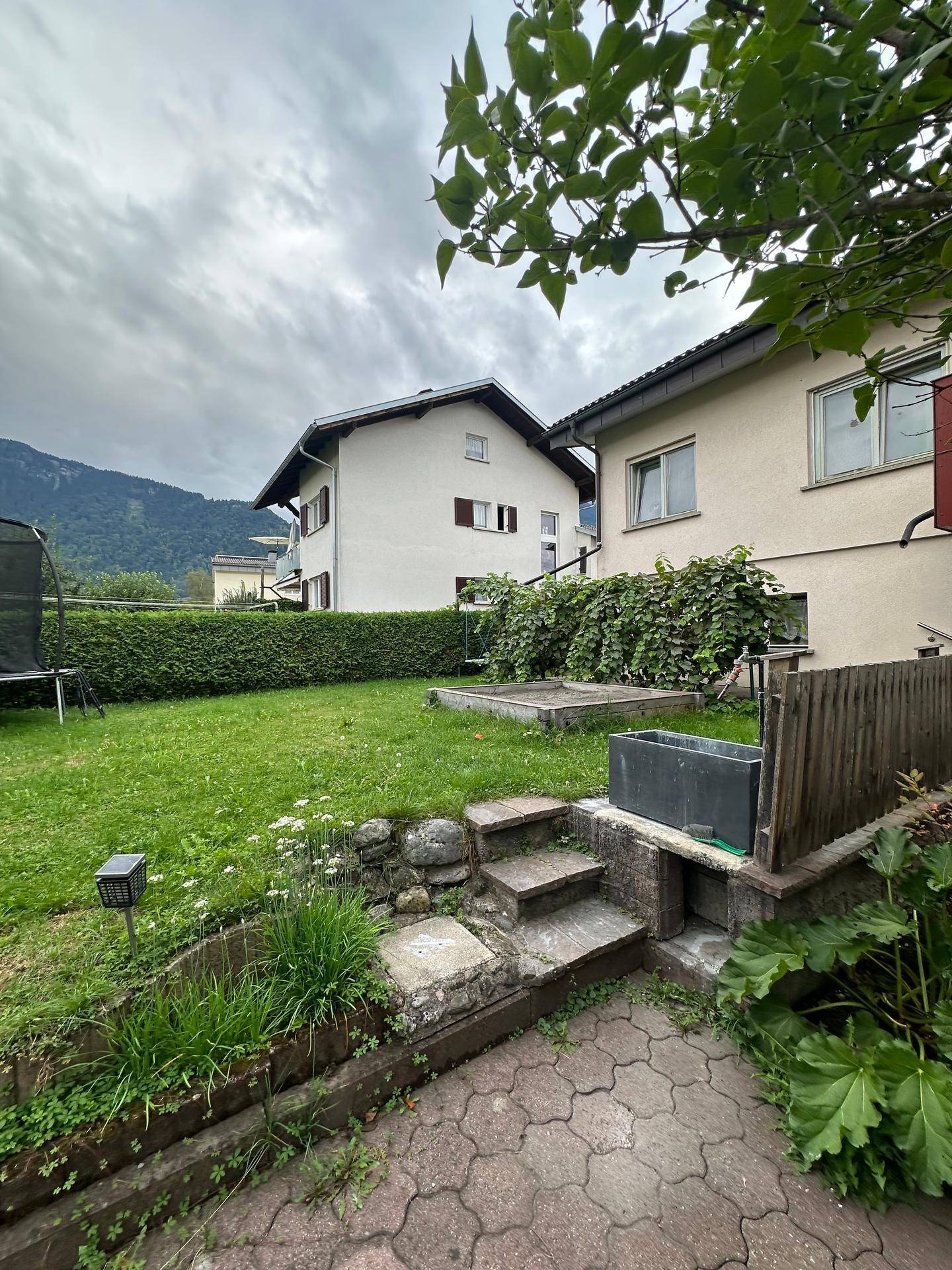 Gartenbereich mit gepflastertem Weg, Steinstufen, Rasenfläche und Blick auf umliegende Häuser und Berge.