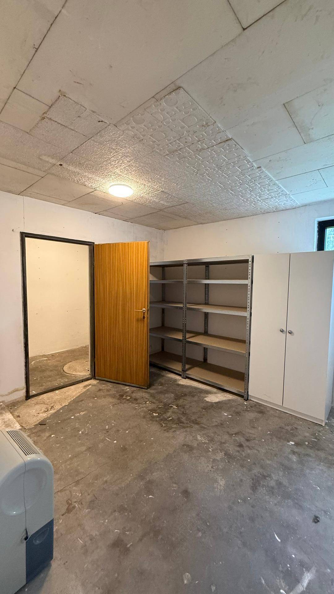 Unfertiger Lagerraum im Keller mit Betonboden, Metallregalen und einem einfachen Holzschrank.