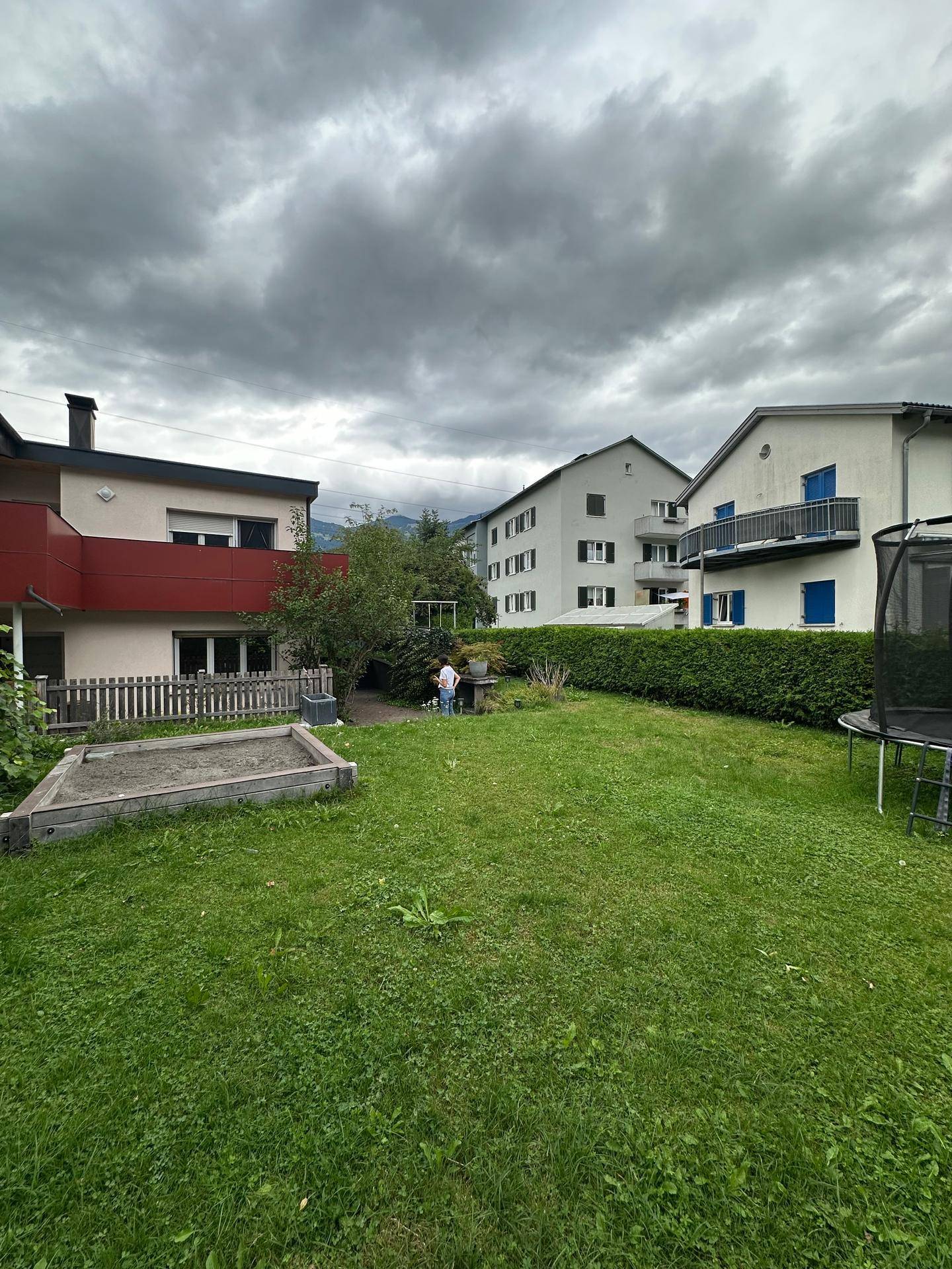 Großer grüner Garten mit Rasenfläche, Sandkasten und Trampolin, umgeben von Wohngebäuden und Bergen im Hintergrund.