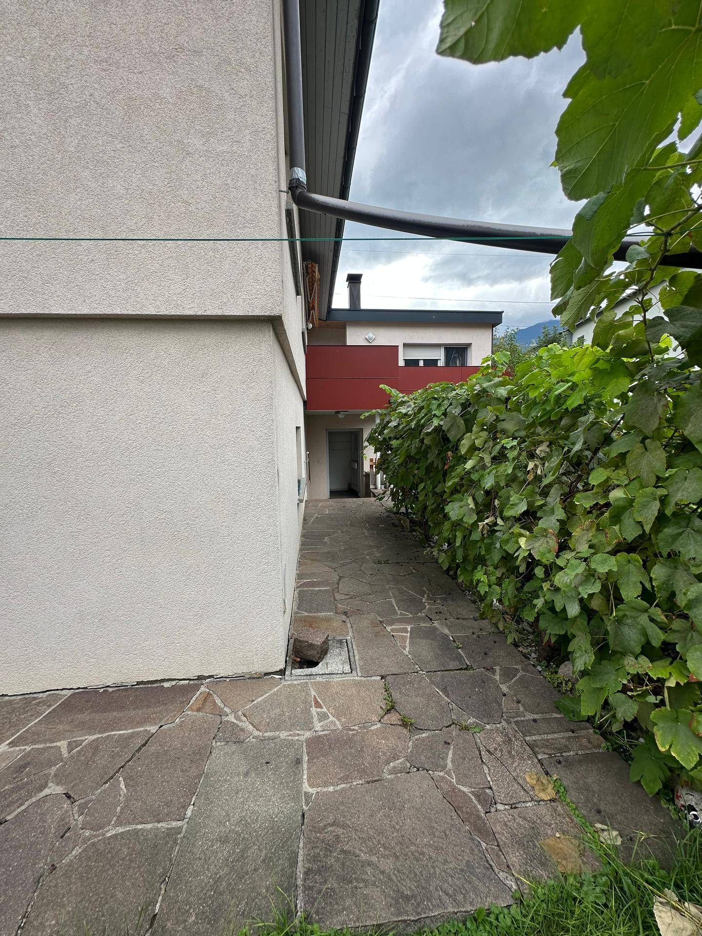 Gepflasterter Weg entlang der Hausfassade mit üppiger Begrünung und Blick auf die Berge.