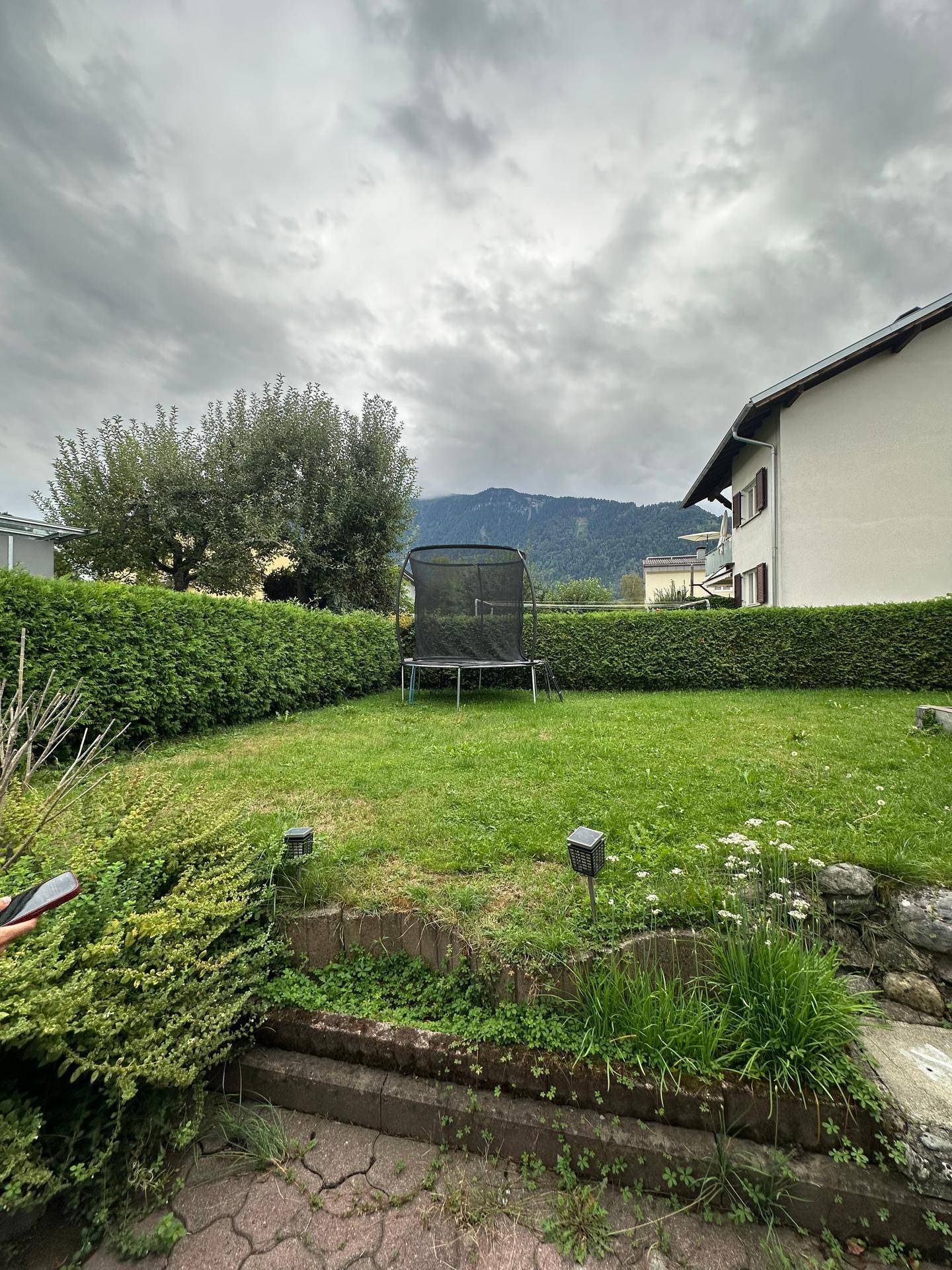 Weitläufiger Rasengarten mit Trampolin, umgeben von einer dichten Hecke und einem malerischen Bergblick.