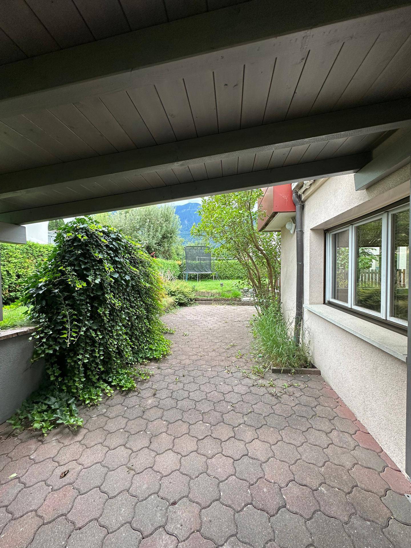 Gepflasterter Gartenweg neben dem Haus mit üppigem Grün und Trampolin im Hintergrund.