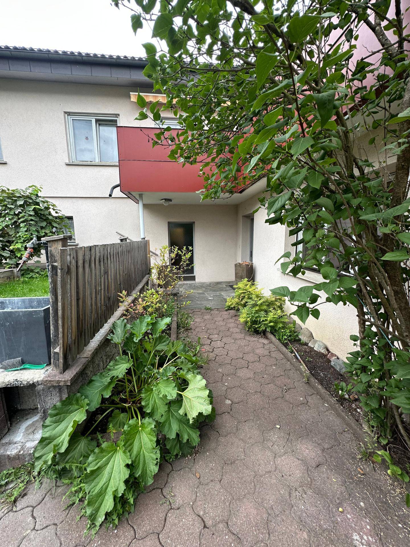 Gepflasterter Zugangsweg zu einem Wohngebäude mit grüner Bepflanzung und einem kleinen Balkon im Hintergrund.