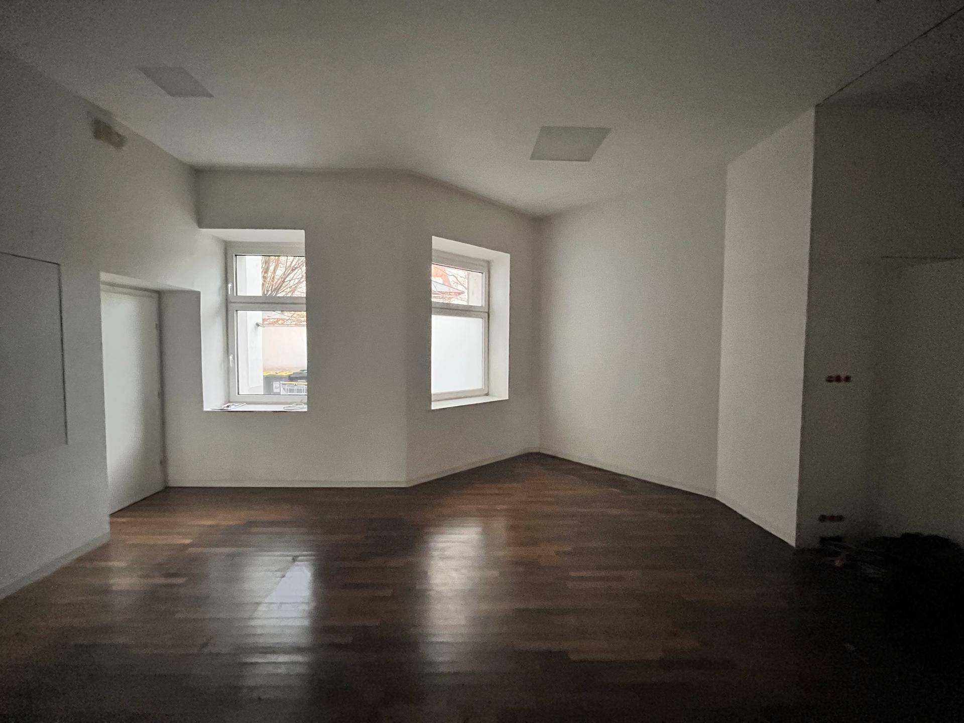 Heller Raum mit dunklem Holzboden und zwei Fenstern, die viel Tageslicht hereinlassen.