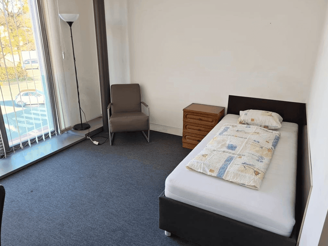 Einfach eingerichtetes Schlafzimmer mit Einzelbett, Nachttisch und großem Fenster.