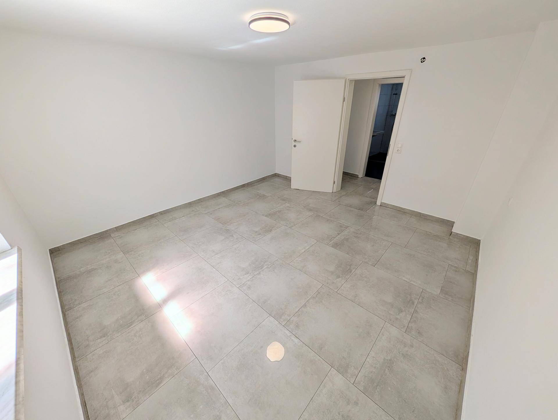 Geräumiger, leerer Raum mit Fliesenboden und offener Tür, die zu einem weiteren Zimmer führt.