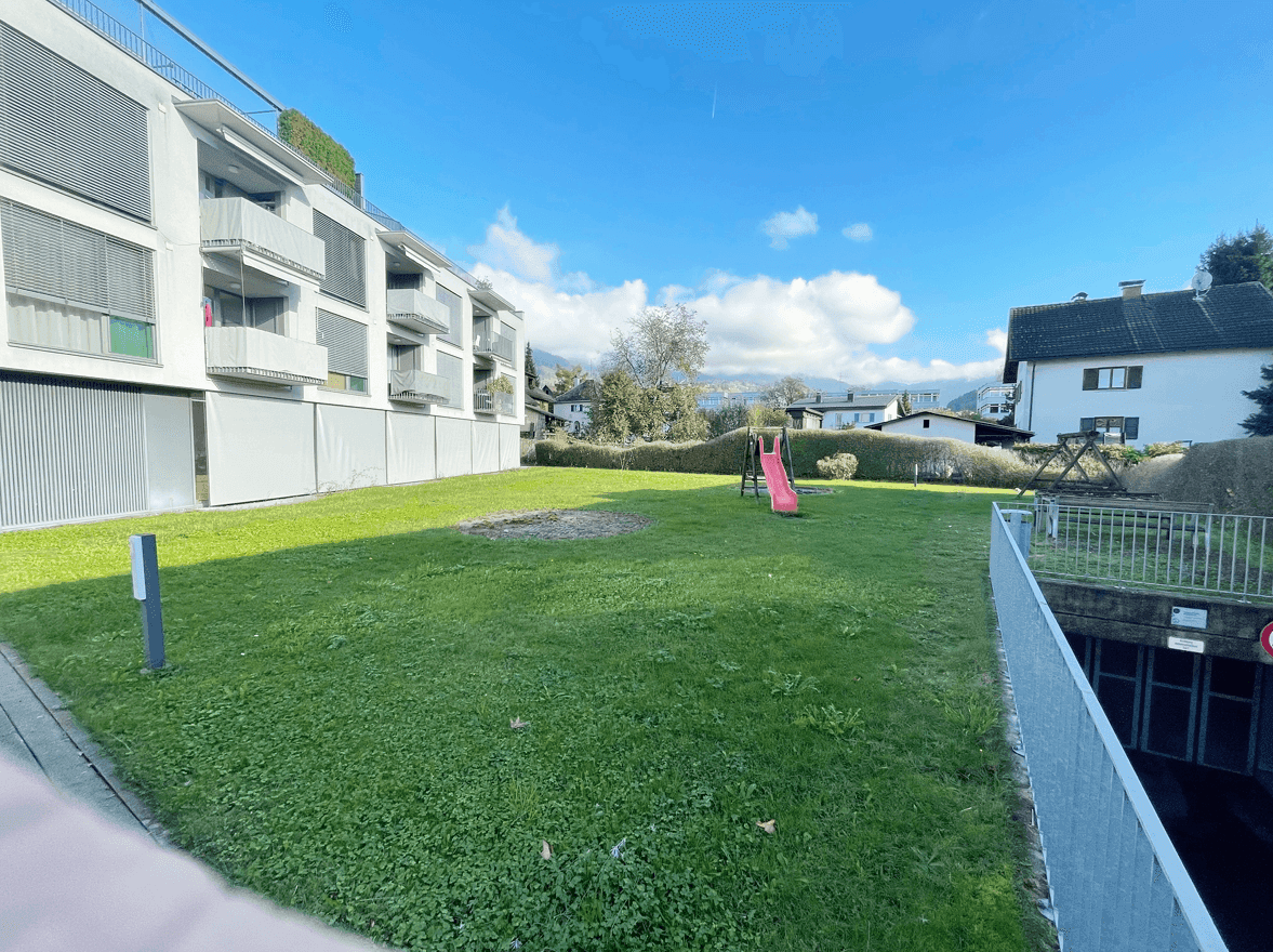 Gepflegter Gemeinschaftsgarten mit Rasenfläche, Kinderrutsche und Blick auf den See im Hintergrund.
