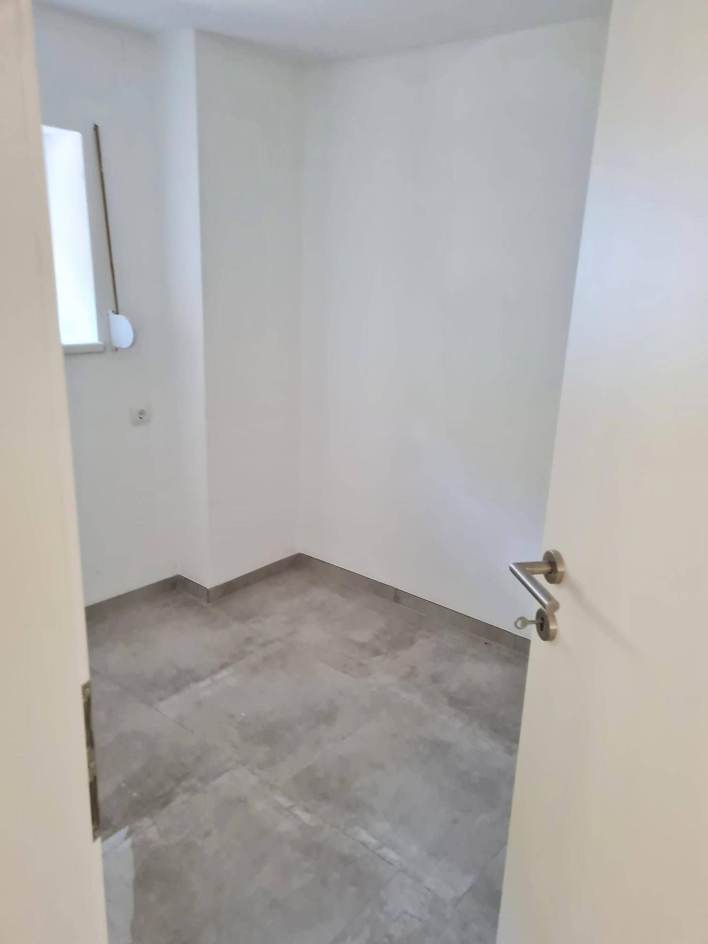 Leeres Zimmer mit Fliesenboden und kleinem Fenster, ideal als Kinder- oder Arbeitszimmer.