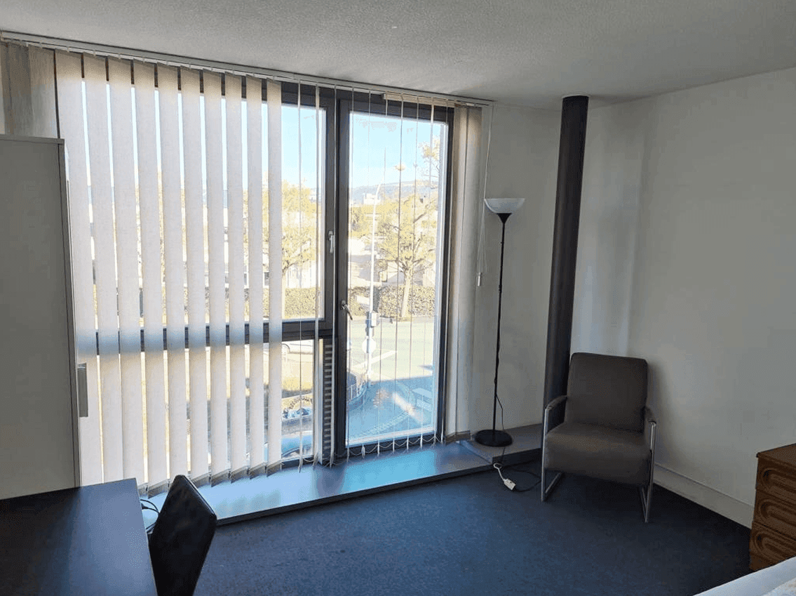 Helles Schlafzimmer mit großem Fenster, vertikalen Jalousien und Blick auf die Straße.