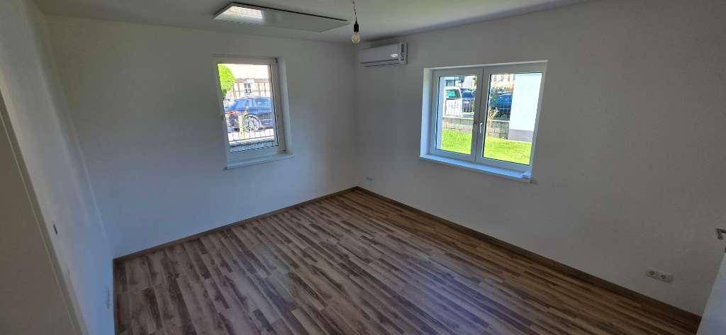 Leeres Zimmer mit Holzboden, zwei Fenstern und einer Klimaanlage an der Wand.