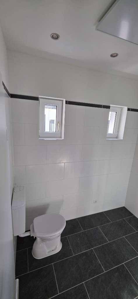 Modernes WC mit Wandtoilette, weißen Wandfliesen und dunklem Fliesenboden.