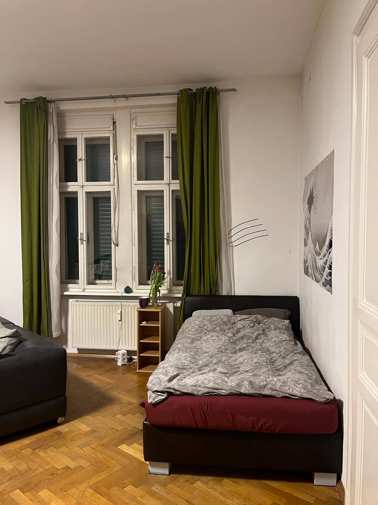 Schlafbereich mit Einzelbett, Parkettboden und grünen Vorhängen an den Fenstern.