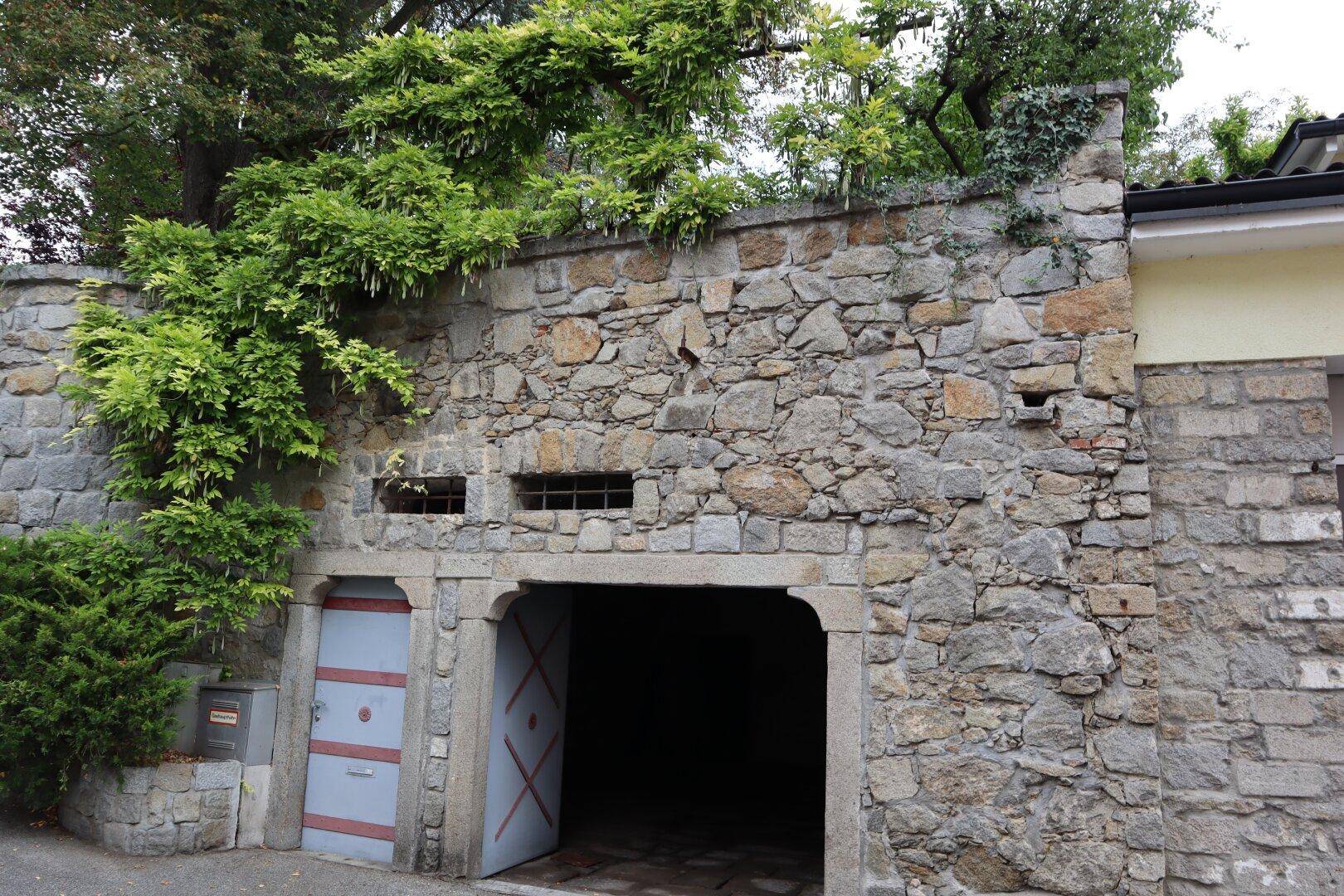 Rustikale Steinmauer mit offenem Torbogen, der zu einem Keller oder einer Garage führt.