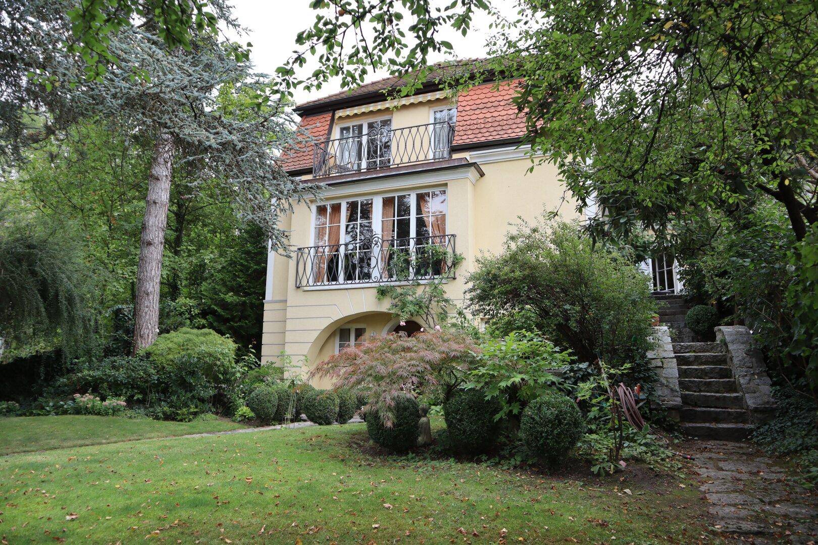 Charmantes Haus mit Mansarddach und gepflegtem Vorgarten umgeben von Bäumen.