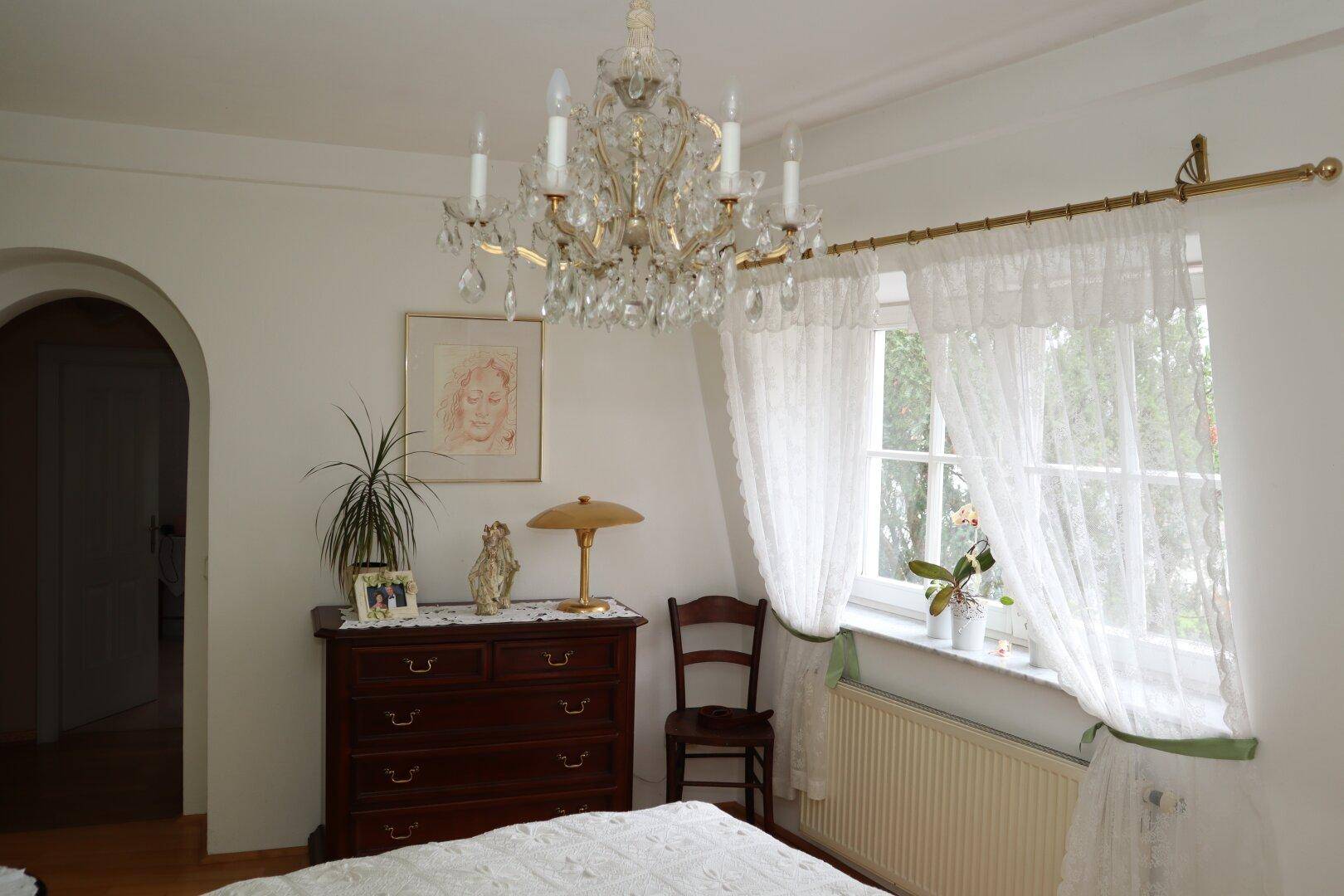 Schlafzimmer mit Kronleuchter, Holzkommode und Fenster mit Spitzenvorhängen.