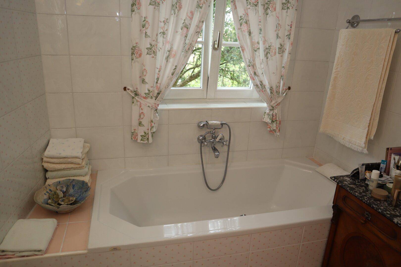 Badezimmer mit Badewanne, Fliesen und Fenster mit floralen Vorhängen.