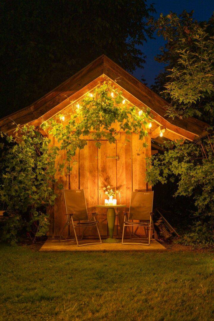 Romantisch beleuchtetes Gartenhaus mit Lichterkette und kleiner Sitzgelegenheit im nächtlichen Garten.