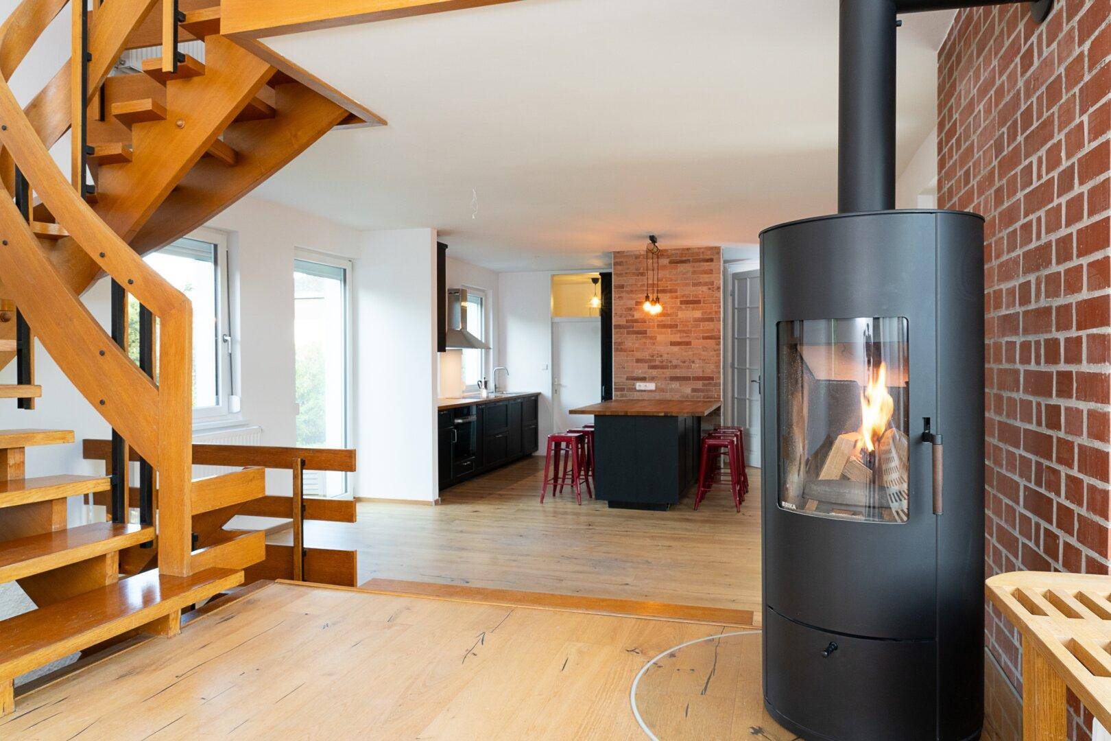 Offener Wohnbereich mit Holztreppe, modernem Kaminofen und integrierter Küche mit Backsteinwand.