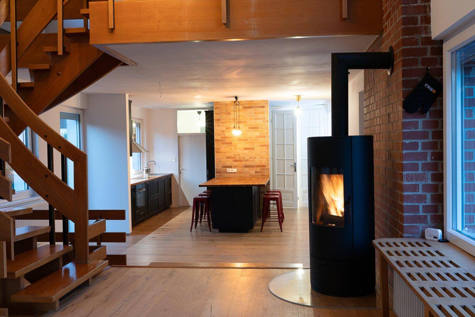 Offener Wohnbereich mit Holztreppe, Kaminofen und integrierter Küche mit Backsteinwand und roten Hockern.