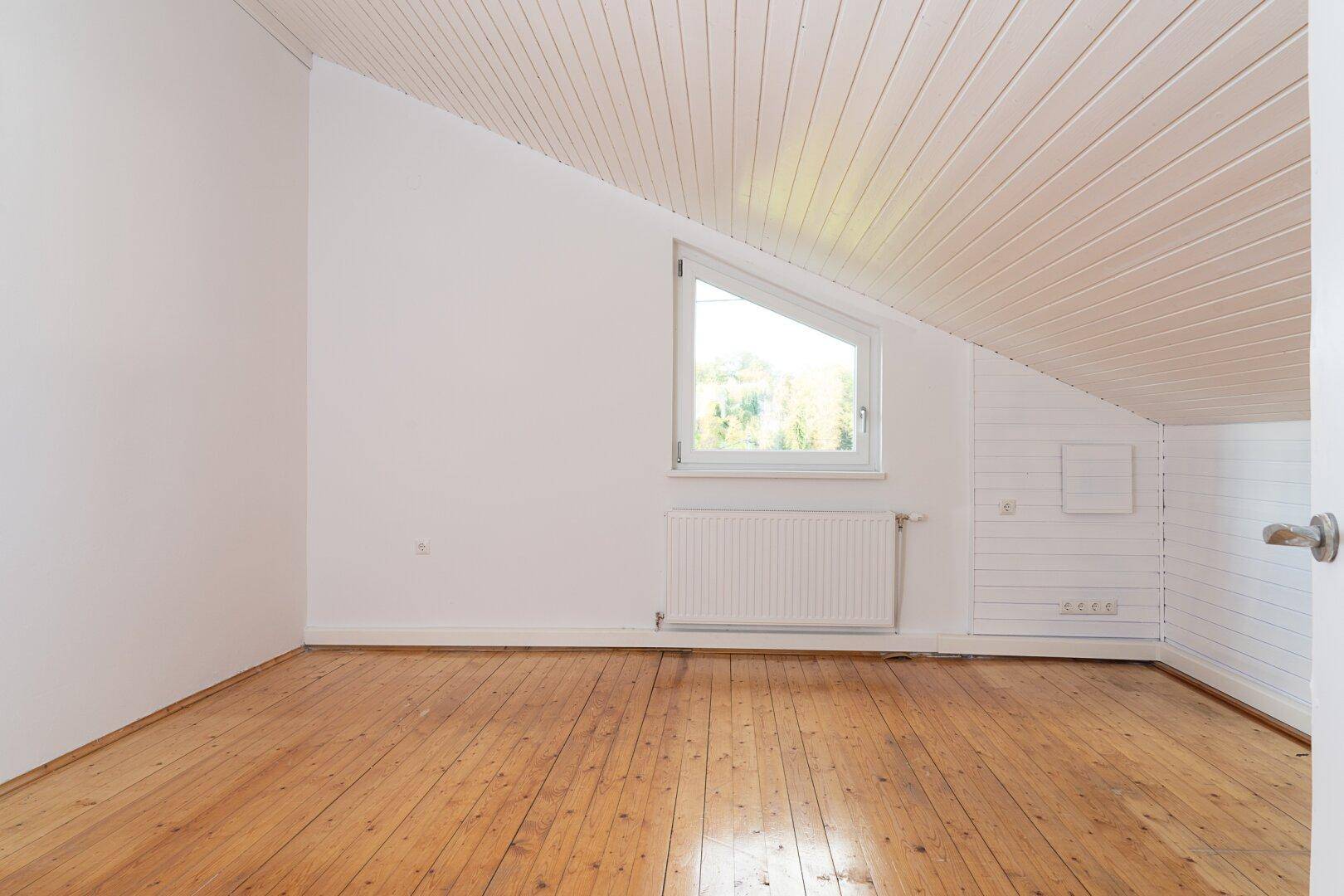 Helles Dachzimmer mit Holzdeckenverkleidung, schrägen Wänden und Holzdielenboden.