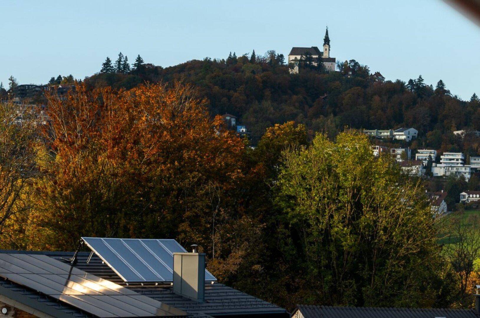 Herbstliche Landschaft mit einer Kirche auf einem Hügel und Solarpaneelen auf einem Dach im Vordergrund.
