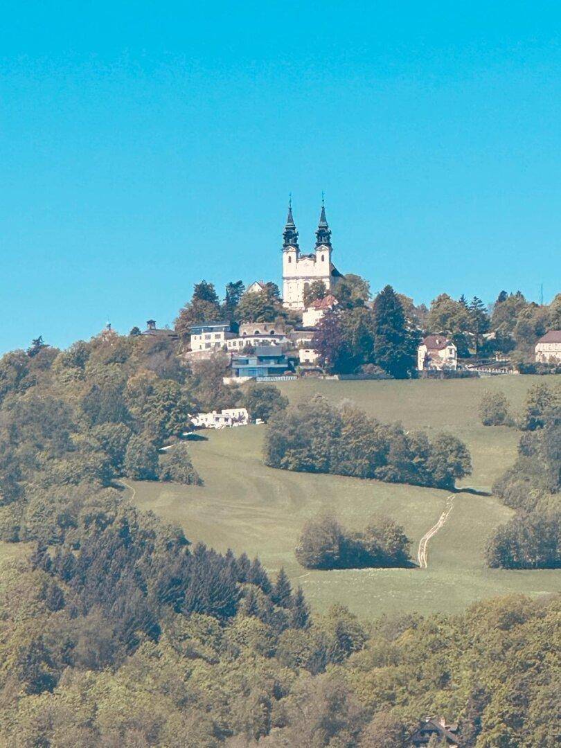 Malerische Landschaft mit einer Kirche auf einem bewaldeten Hügel unter strahlend blauem Himmel.