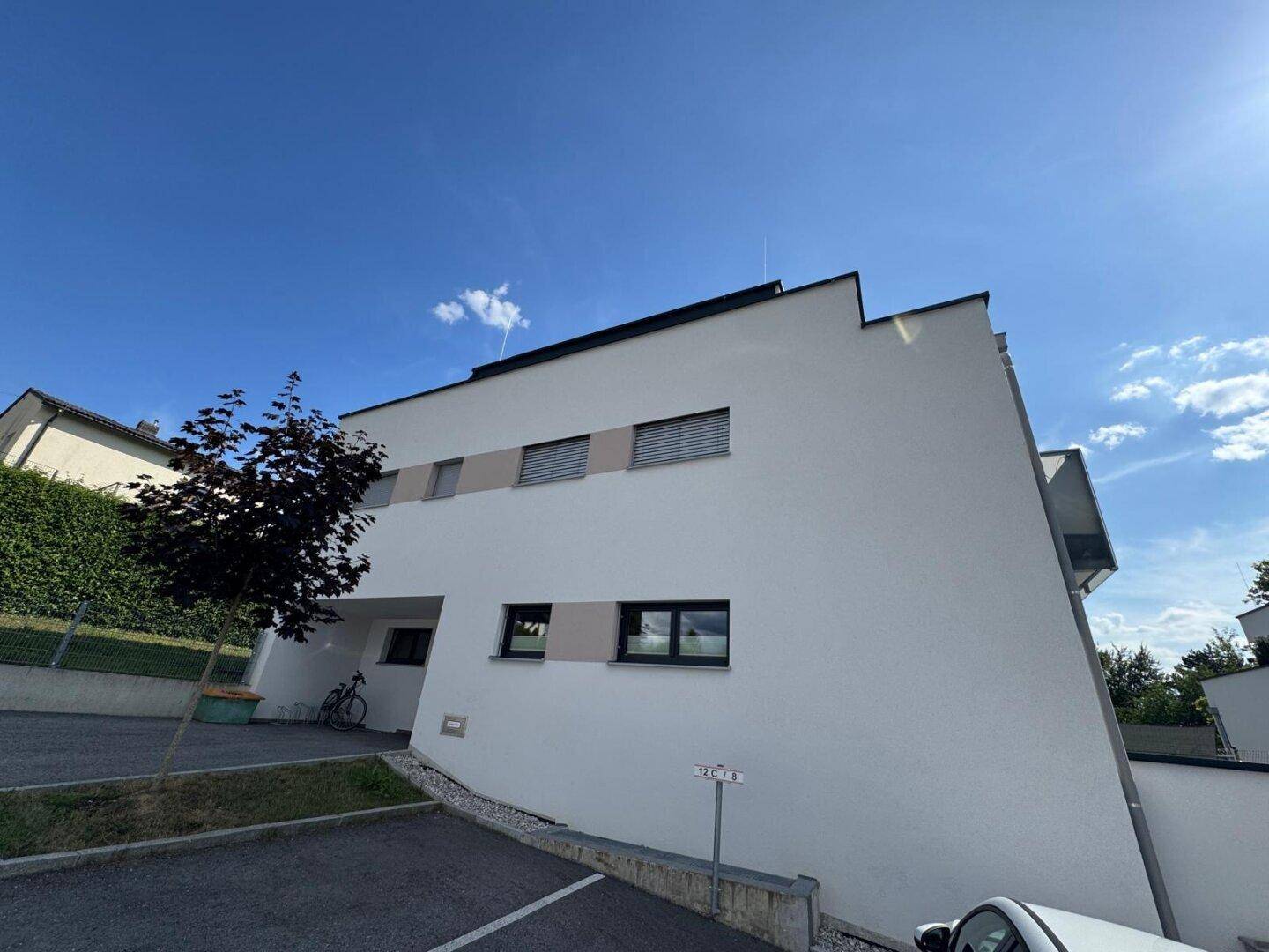 Außenansicht des modernen Wohnhauses mit weißer Fassade, Parkplatz und klarem Himmel.