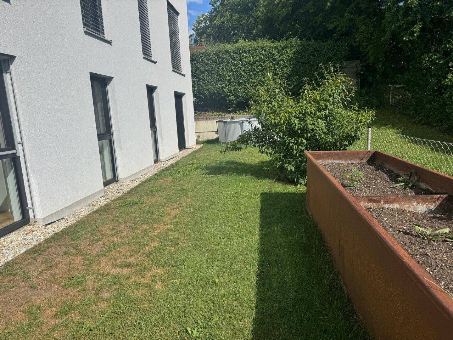 Schmaler Gartenbereich neben dem Gebäude mit Rasenfläche, Hochbeet und Hecke.