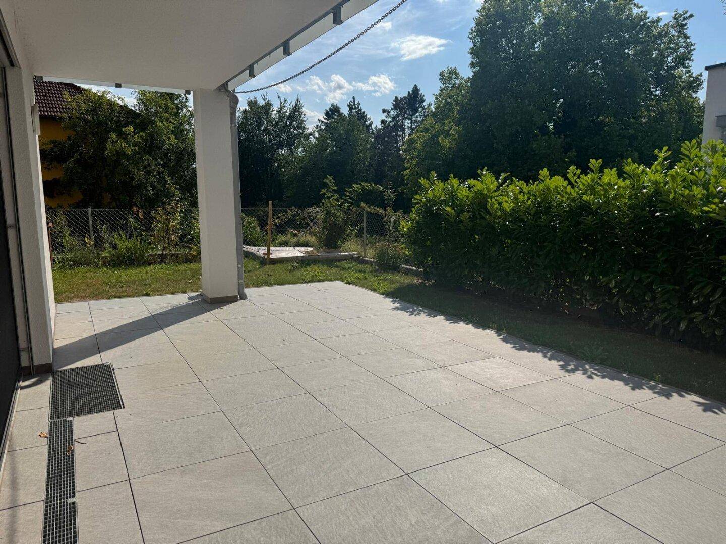 Helle geflieste Terrasse mit Überdachung, angrenzendem Rasen und grüner Hecke.