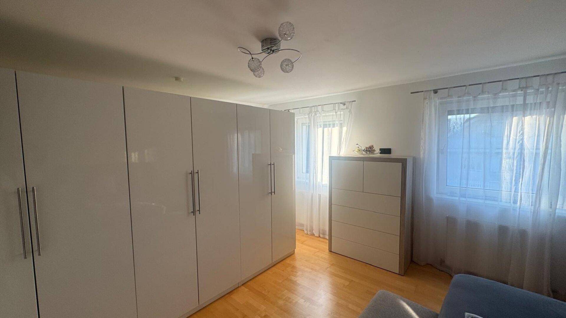 Geräumiges Schlafzimmer mit glänzenden weißen Kleiderschränken, Holzboden und zwei Fenstern.
