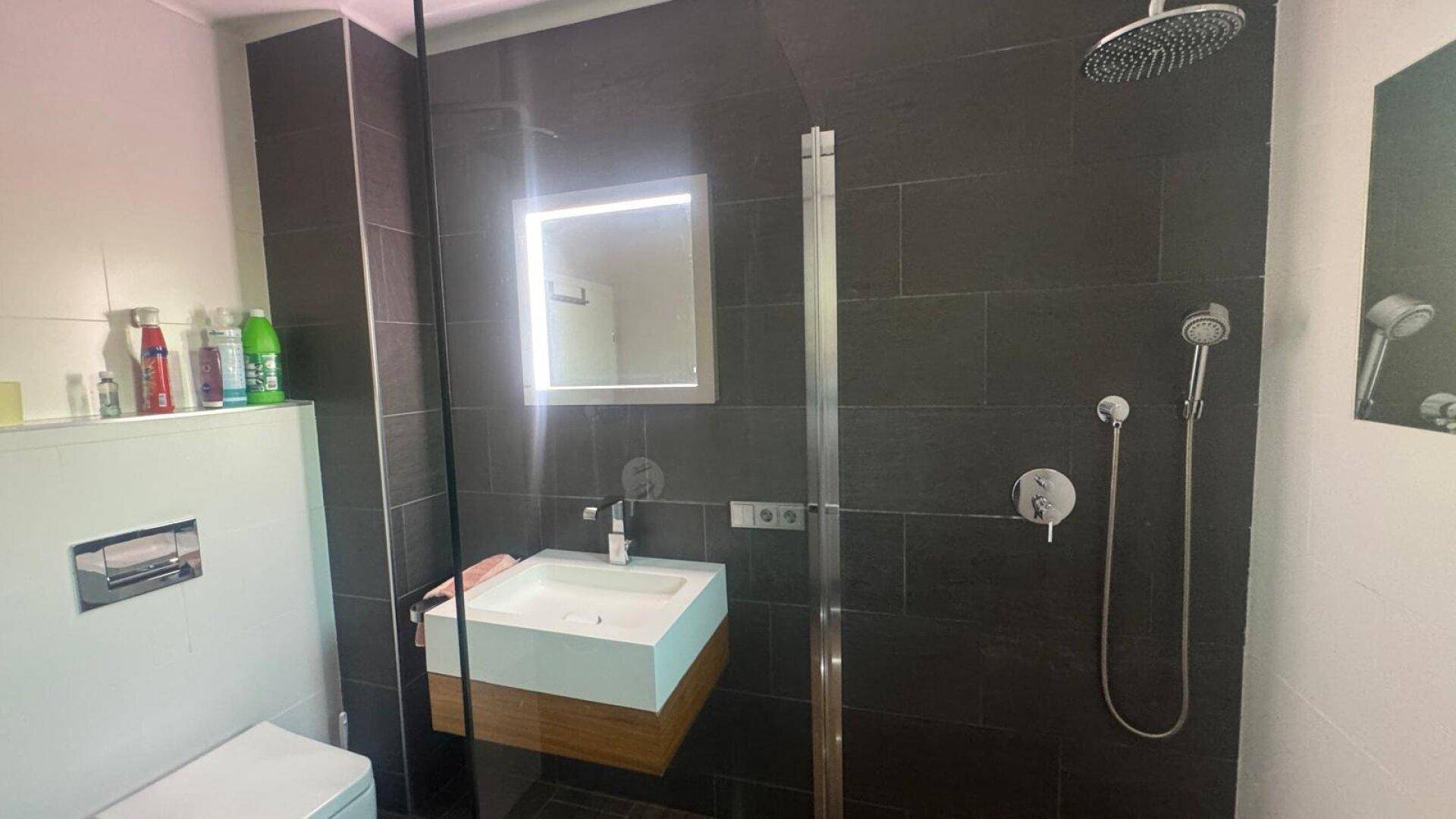 Modernes Badezimmer mit großer begehbarer Dusche, Waschtisch und dunklen Wandfliesen.