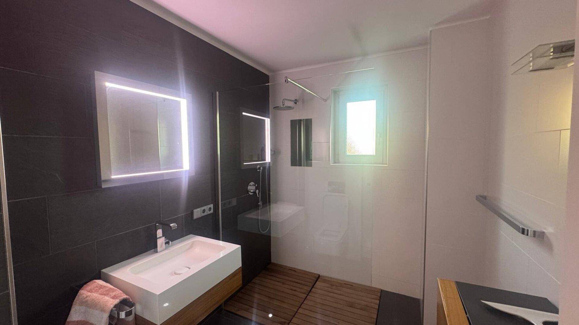 Modernes Badezimmer mit begehbarer Dusche, beleuchtetem Spiegel und dunklen Fliesen.