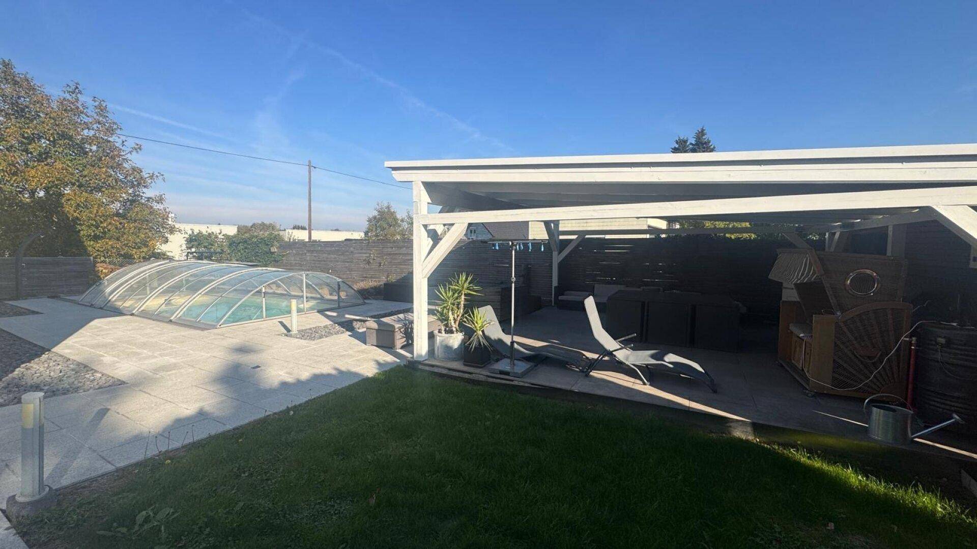 Außenbereich mit überdachtem Pool, gepflasterter Terrasse und einem Carport im Hintergrund.