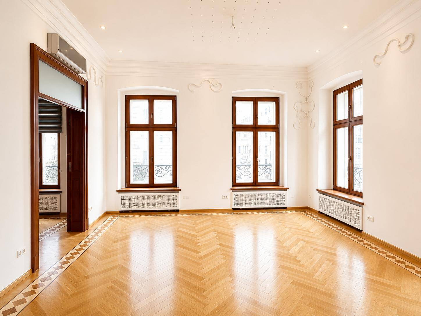 Heller, leerer Raum mit Fischgrätparkett, hohen Decken und großen Fenstern mit Blick nach draußen.