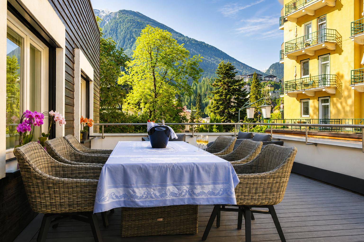 Geräumige Terrasse mit Esstisch und Korbstühlen, umgeben von grünen Bergen.