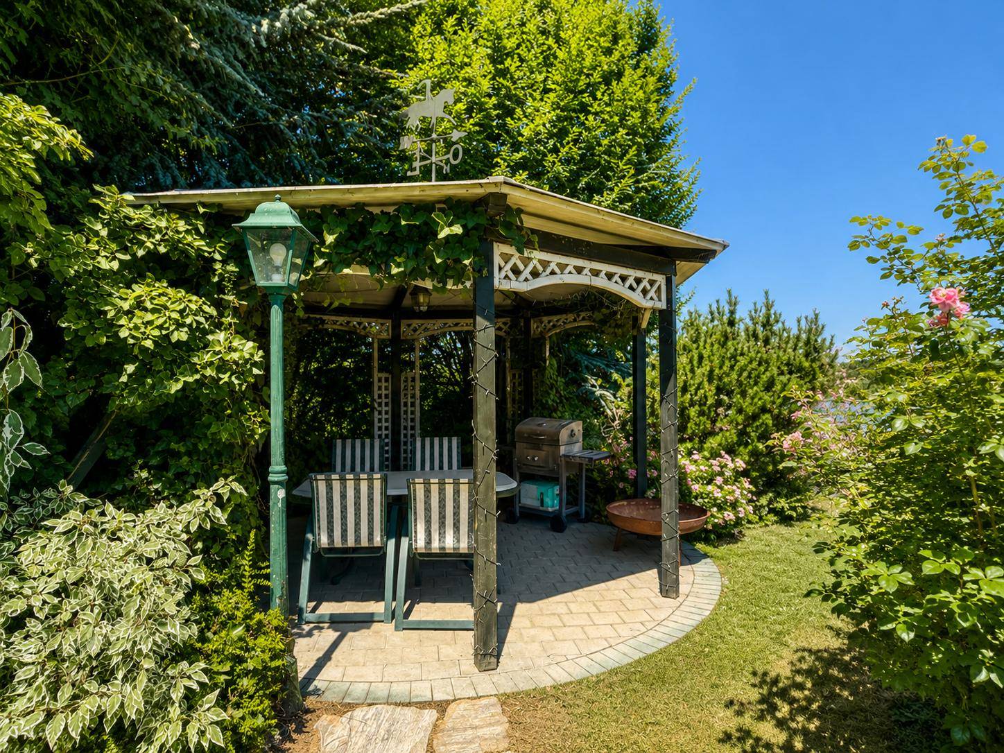 Gemütlicher Gartenpavillon mit Sitzgelegenheiten und Grillplatz, umgeben von üppigem Grün.