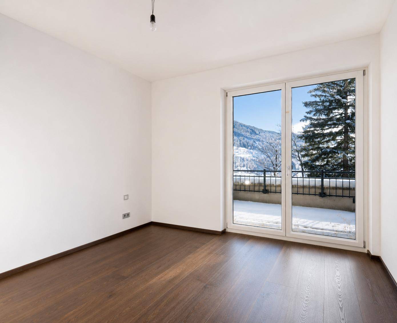 Helles Zimmer mit dunklem Holzboden und großer Schiebetür, die Zugang zu einem Balkon mit Bergblick bietet.