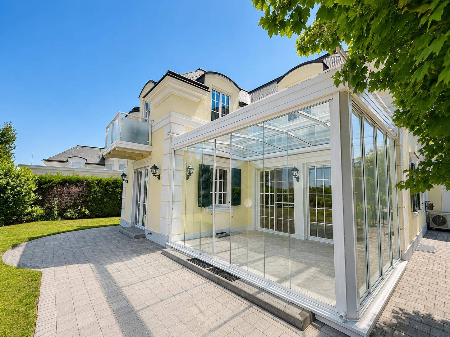 Elegantes Haus mit großem Wintergarten und gepflasterter Terrasse, umgeben von einem gepflegten Garten.