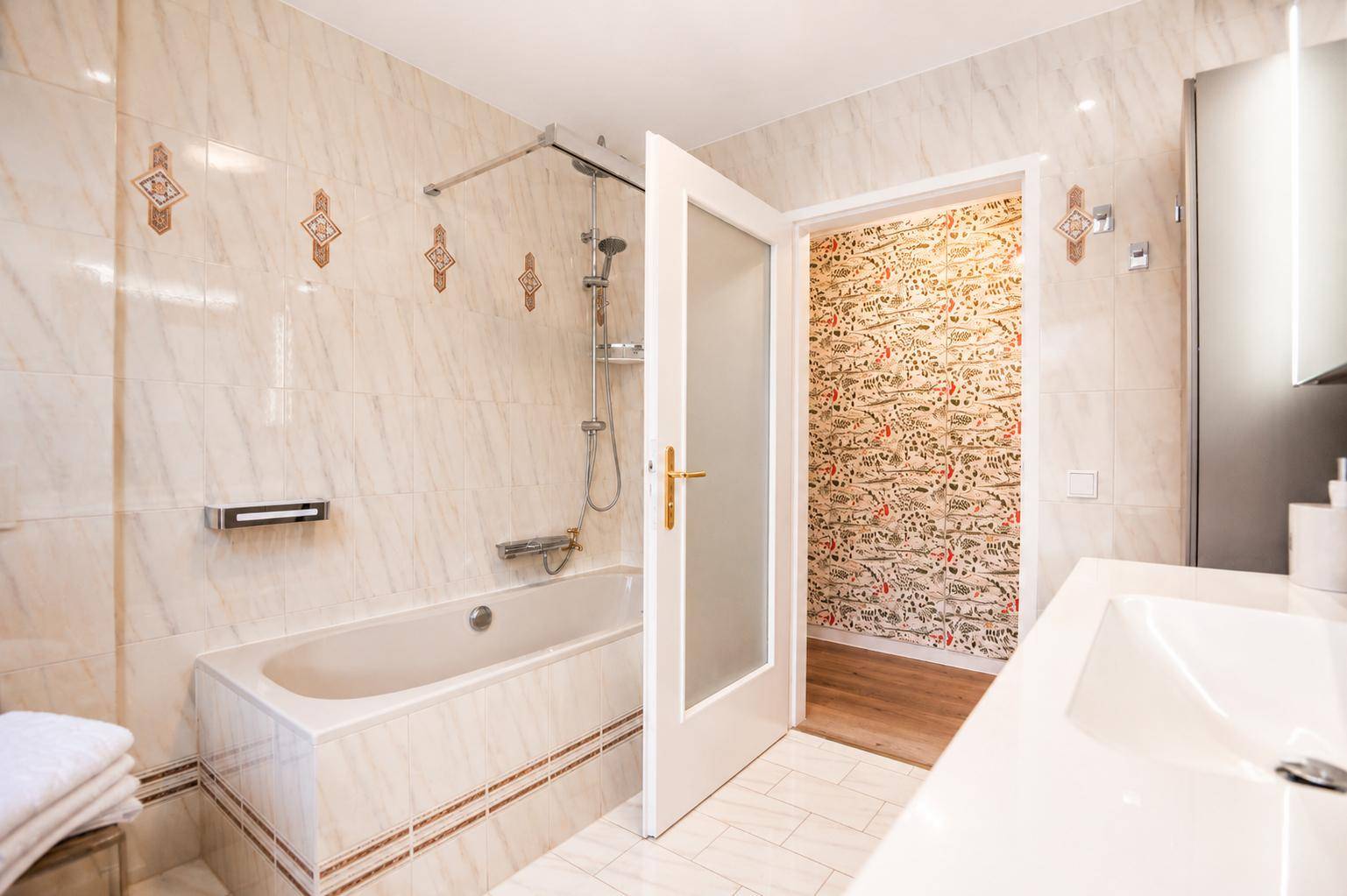 Gefliestes Badezimmer mit Badewanne und Duschvorrichtung, angrenzend an einen Raum mit Tapete.