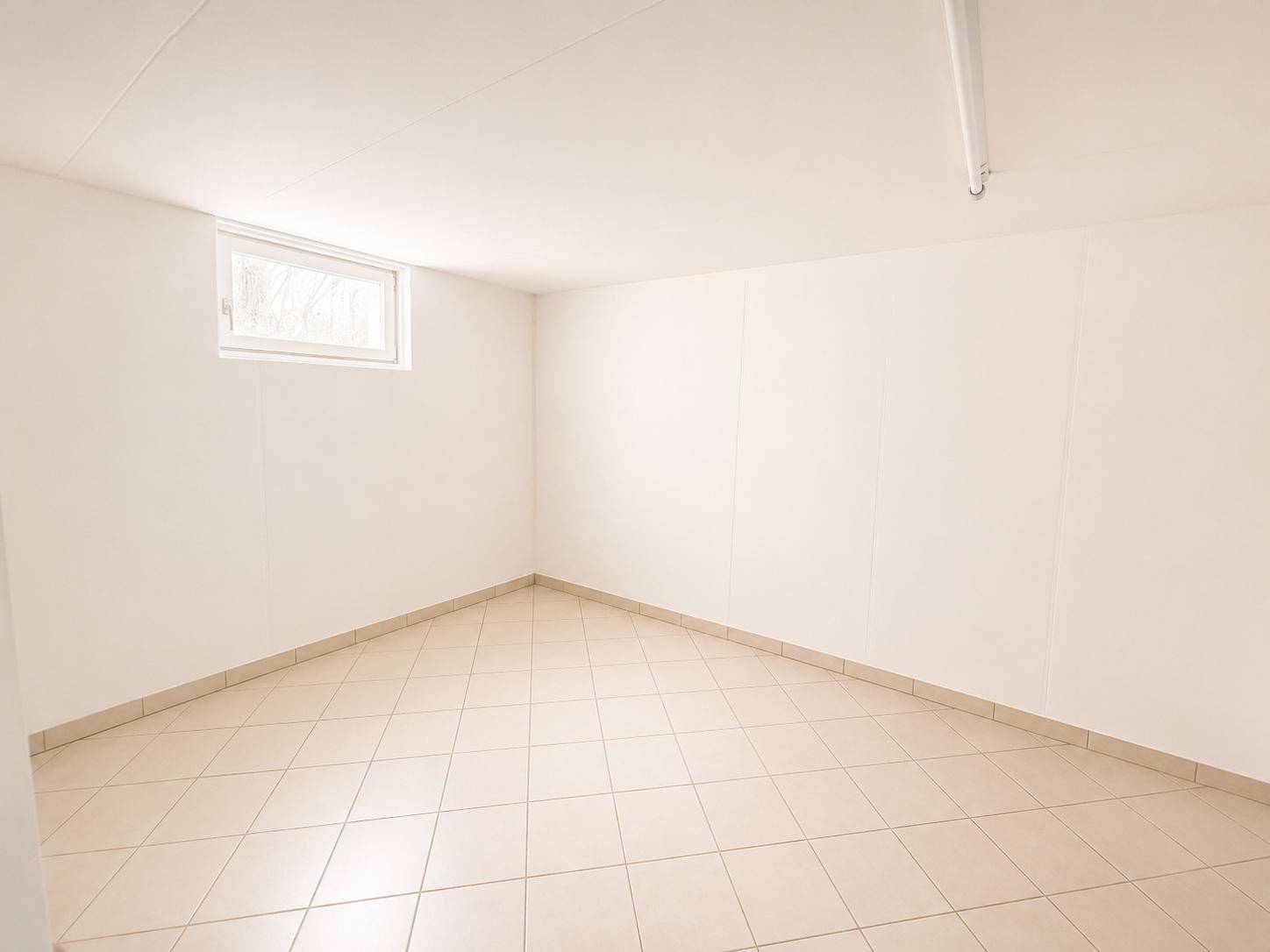 Leerer Kellerraum mit Fliesenboden und kleinem Fenster, ideal als Abstellraum.