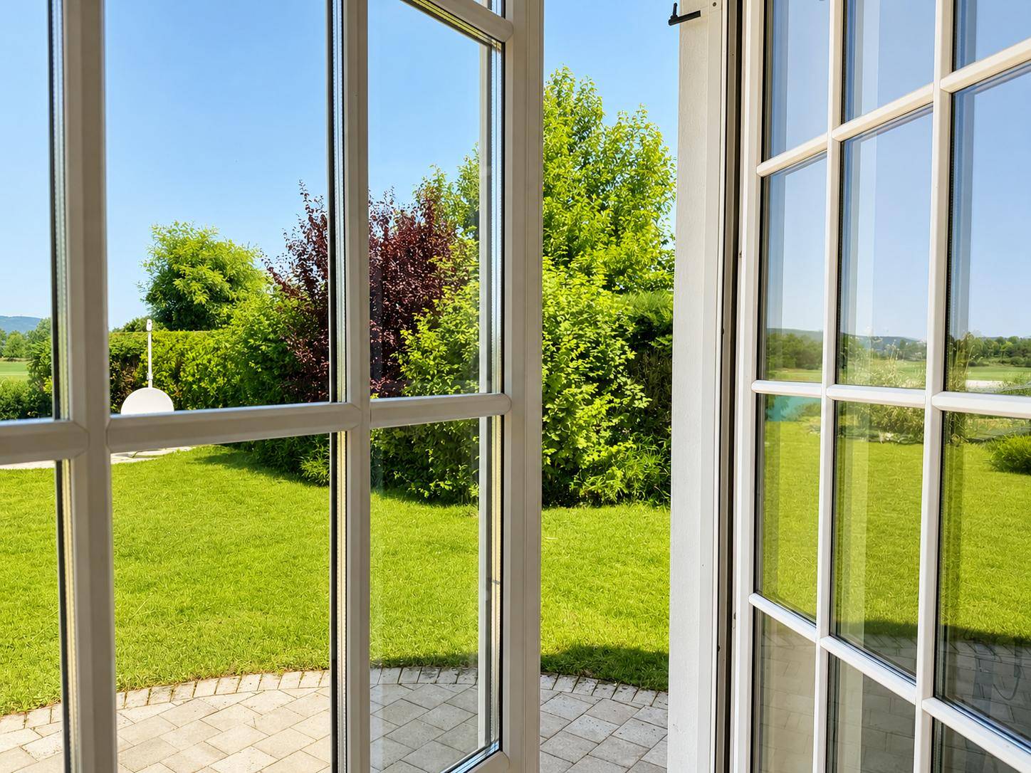 Blick durch ein großes Fenster auf den gepflegten grünen Garten an einem sonnigen Tag.