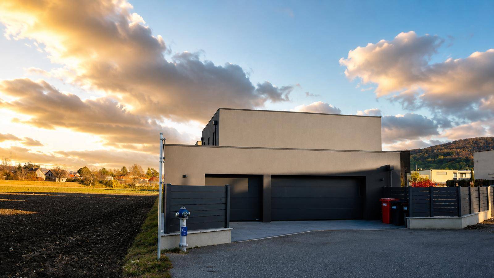 Modernes Haus mit Doppelgarage und Zaun, angrenzend an einen Acker unter einem bewölkten Himmel.