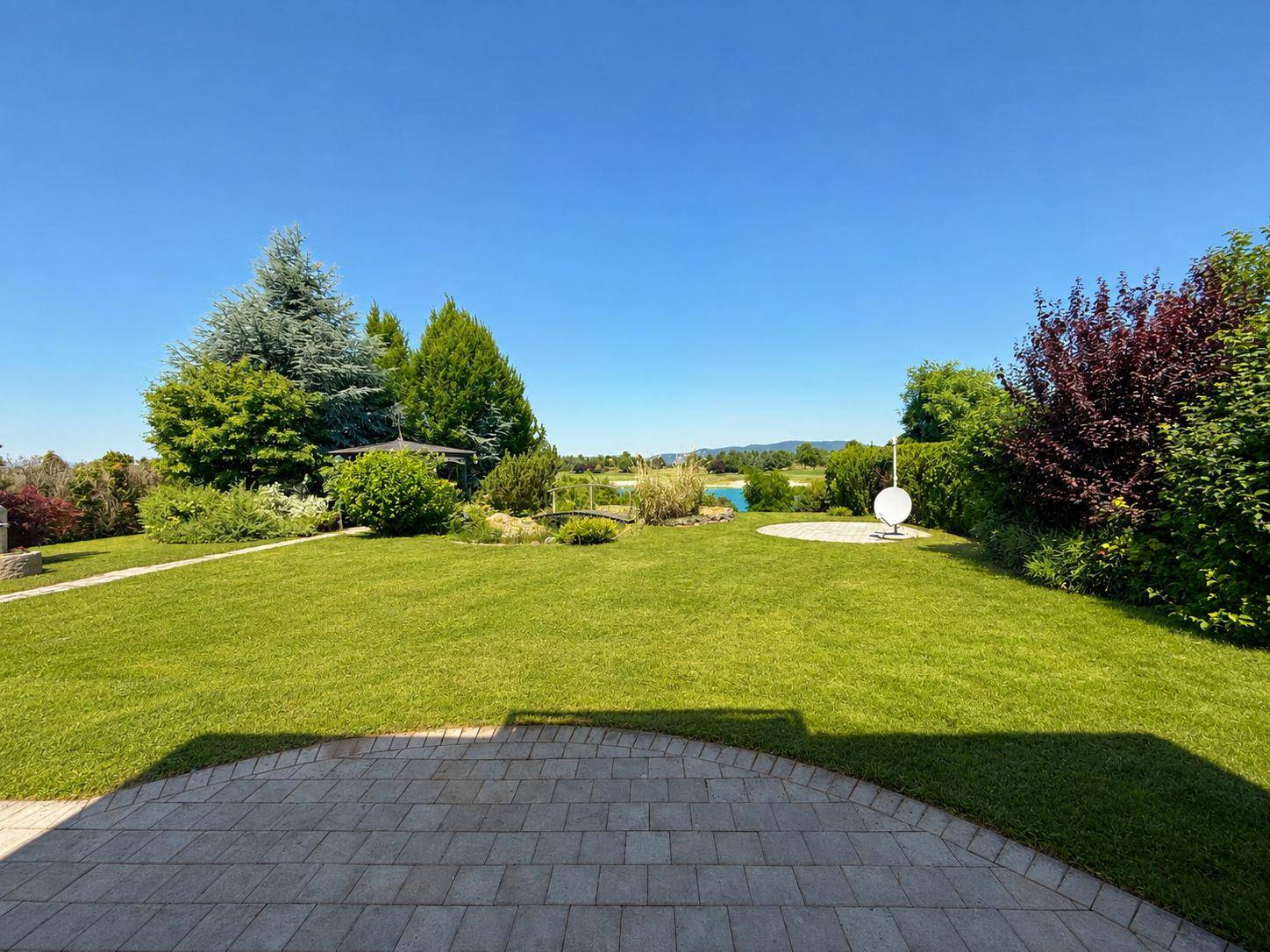 Große Rasenfläche mit gepflasterter Terrasse und weitem Blick auf die umliegende Landschaft und den See.