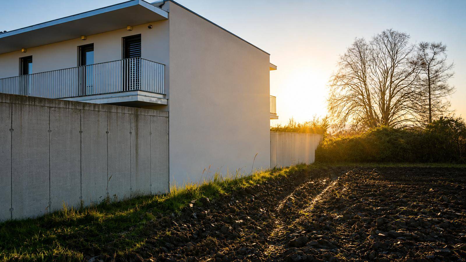 Modernes Gebäude mit Balkonen und einer Betonmauer, angrenzend an einen Acker bei Sonnenuntergang.