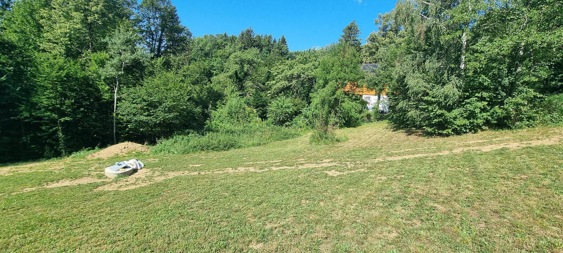 Grüne Wiese mit Waldrand, Erdhaufen und einem Betonschacht unter blauem Himmel.