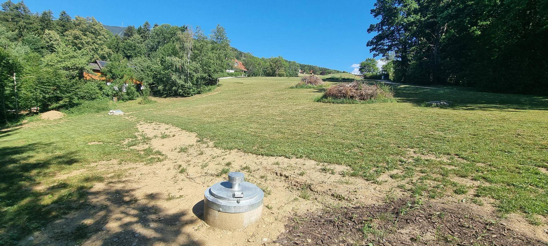 Grüne Wiese mit einem Betonschacht im Vordergrund und Blick auf die hügelige Landschaft.
